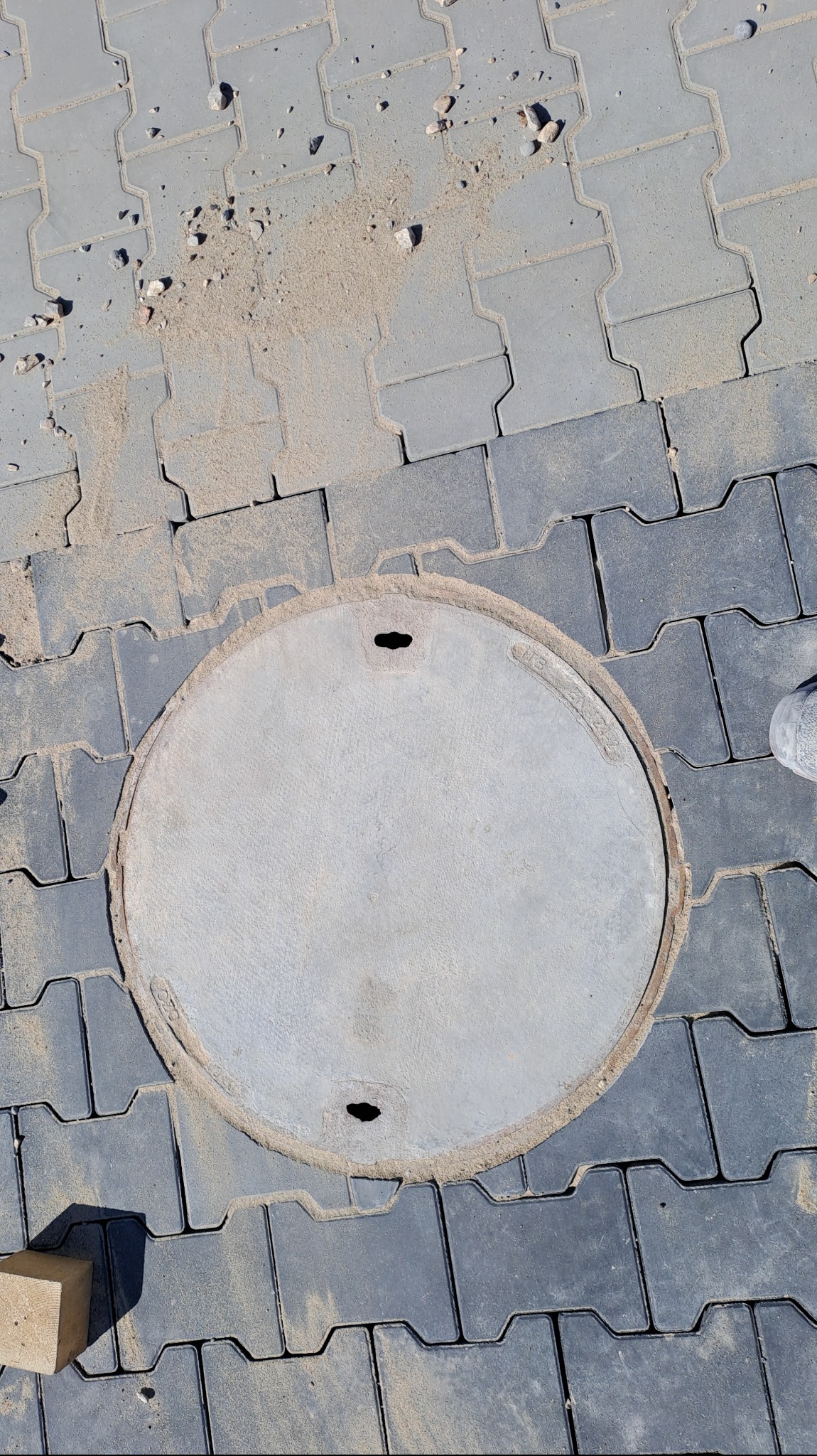 Szare, betonowe płyty chodnikowe ułożone wokół okrągłej studzienki kanalizacyjnej. Widoczne drobne zanieczyszczenia piaskiem i gruzem oraz fragment drewnianego klocka.