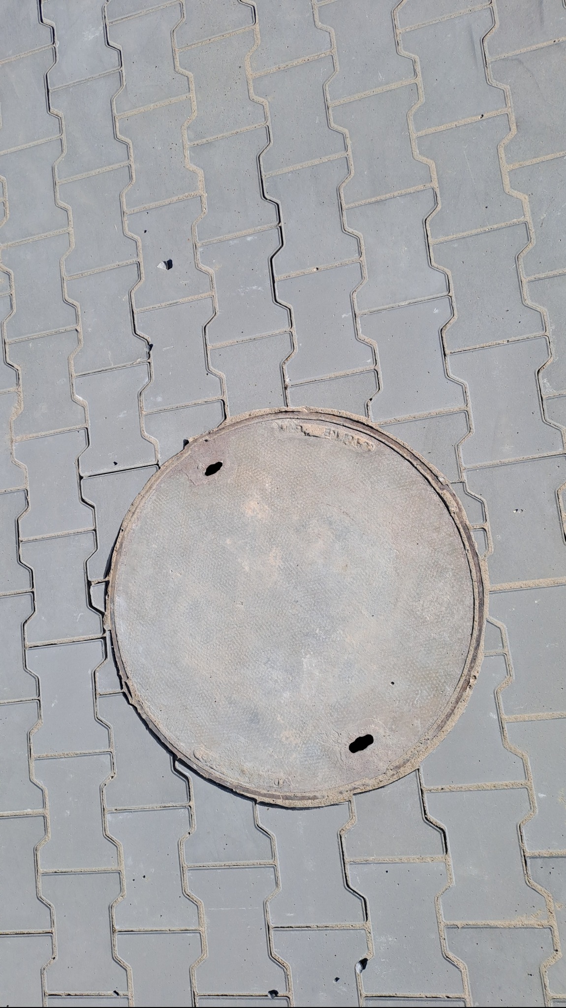 Szara kostka brukowa typu 'I' z okrągłą, żeliwną studzienką kanalizacyjną z dwoma otworami.