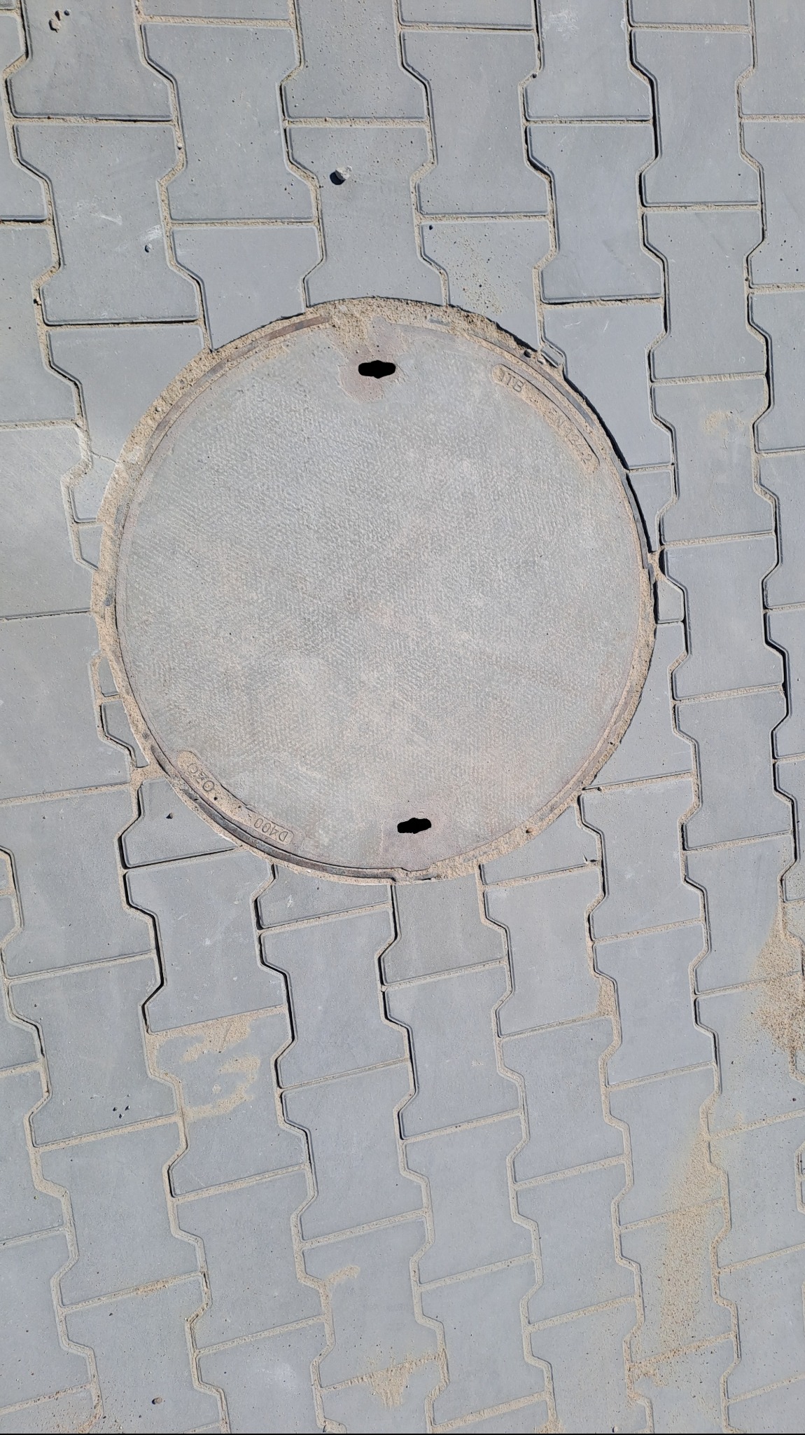Szara kostka brukowa typu I z okrągłą, żeliwną studzienką kanalizacyjną z dwoma otworami na klucz.