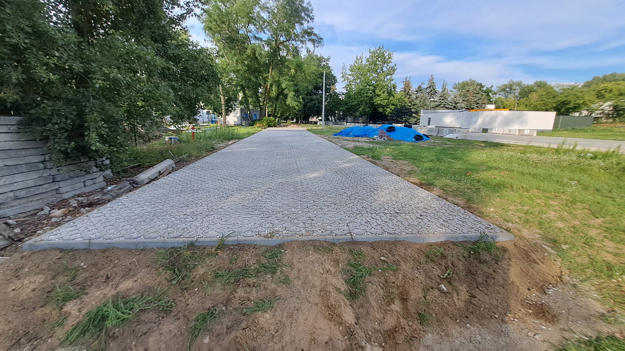 Długa, szara ścieżka z kostki brukowej o nieregularnym wzorze, otoczona ziemią i zielenią. W tle drzewa i budynek z niebieską plandeką.
