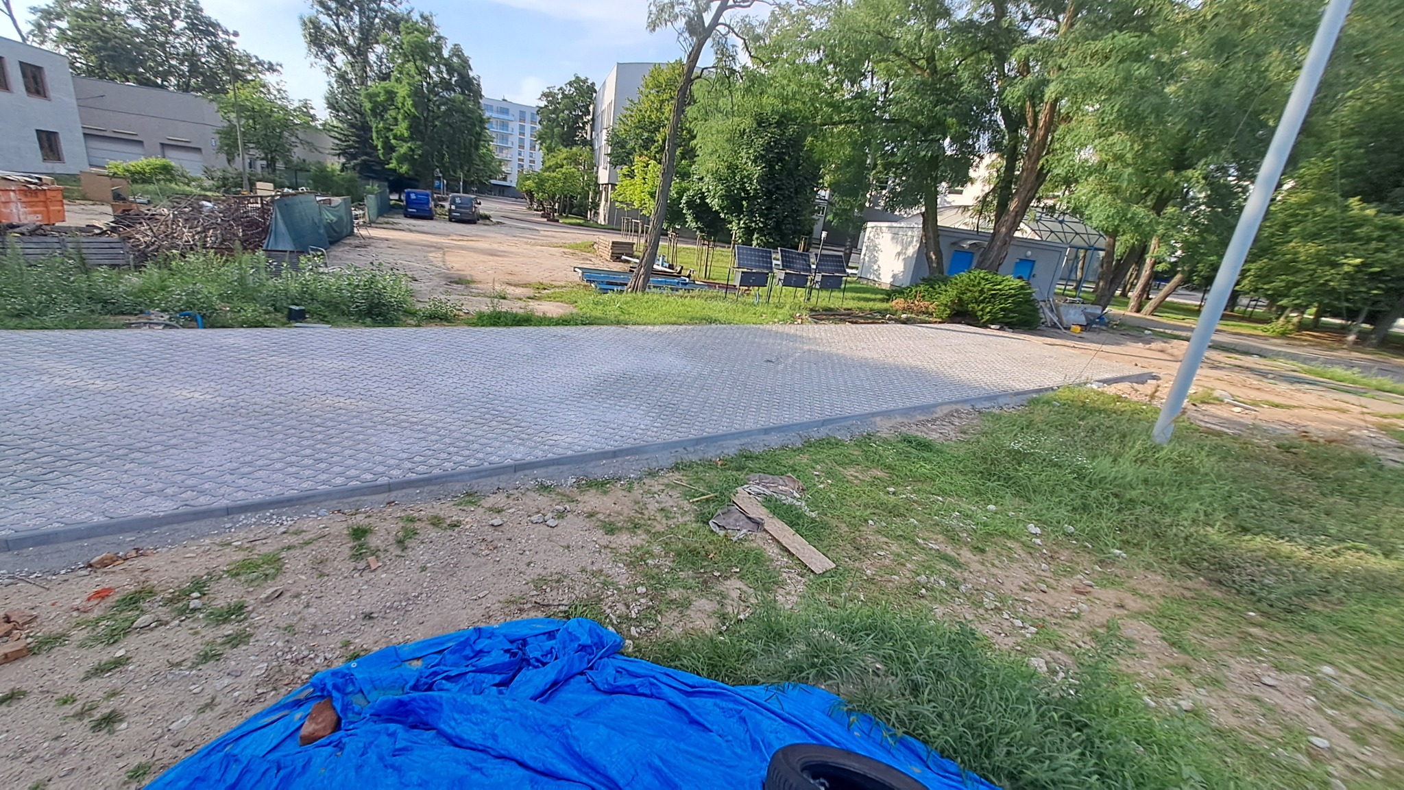 Ułożona szara kostka brukowa na posesji, widok z perspektywy trawnika. W tle budynki i drzewa, a w rogu kadru fragment niebieskiej plandeki.