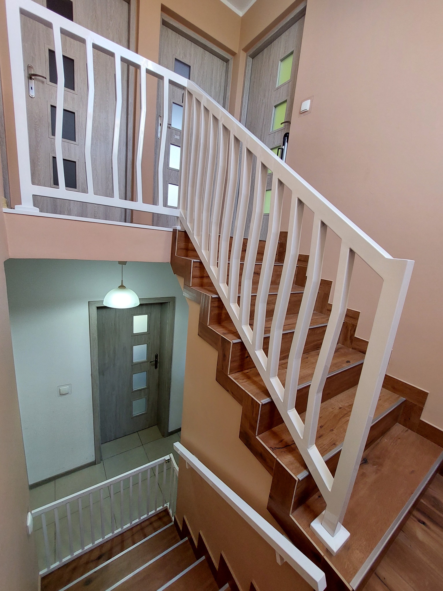 Wewnętrzna klatka schodowa z drewnianymi schodami i białymi, nowoczesnymi balustradami. Widok z góry na dół, ukazujący schody prowadzące na dwa poziomy, z trzema parami drzwi widocznymi w tle.