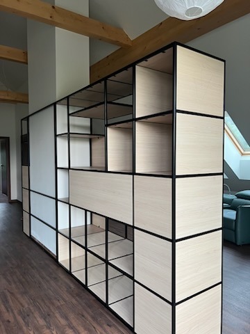 Nowoczesna, modułowa konstrukcja regałowa z czarnych metalowych ram i jasnego drewna w przestronnym wnętrzu z drewnianą podłogą i belkami stropowymi.