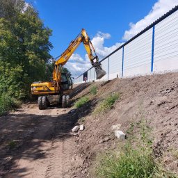 Rafał Grajek RG-EKOPROJEKT - Żółta koparka na nierównym terenie, przygotowująca teren pod budowę, widoczna długa, biała ściana z niebieskimi akcentami w tle.