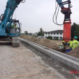 Rafał Grajek RG-EKOPROJEKT - Układanie krawężnika drogowego z użyciem specjalistycznej maszyny w wykopie; w tle koparka i budynek.