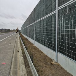 Rafał Grajek RG-EKOPROJEKT - Długa, akustyczna ściana z metalową siatką i betonową podstawą wzdłuż drogi, z koparką w tle i pochmurnym niebem.