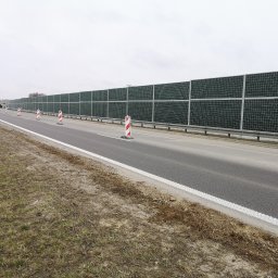 Rafał Grajek RG-EKOPROJEKT - Długa bariera akustyczna z zielonymi panelami wzdłuż drogi, z widocznymi pachołkami ostrzegawczymi i sprzętem budowlanym w oddali.