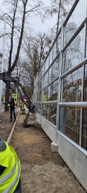 Montaż ekranu akustycznego przy drodze: koparka Mecalac przygotowuje teren pod fundamenty, widoczni pracownicy w odblaskowych kamizelkach, drzewa w tle.
