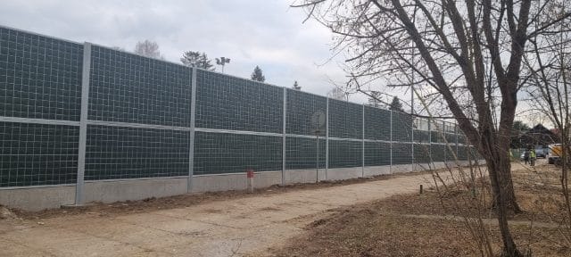 Długa, zielona bariera akustyczna z metalową ramą i betonową podstawą, ustawiona wzdłuż drogi gruntowej, z widocznymi drzewami bez liści i częściowo widocznymi pojazdami w tle.