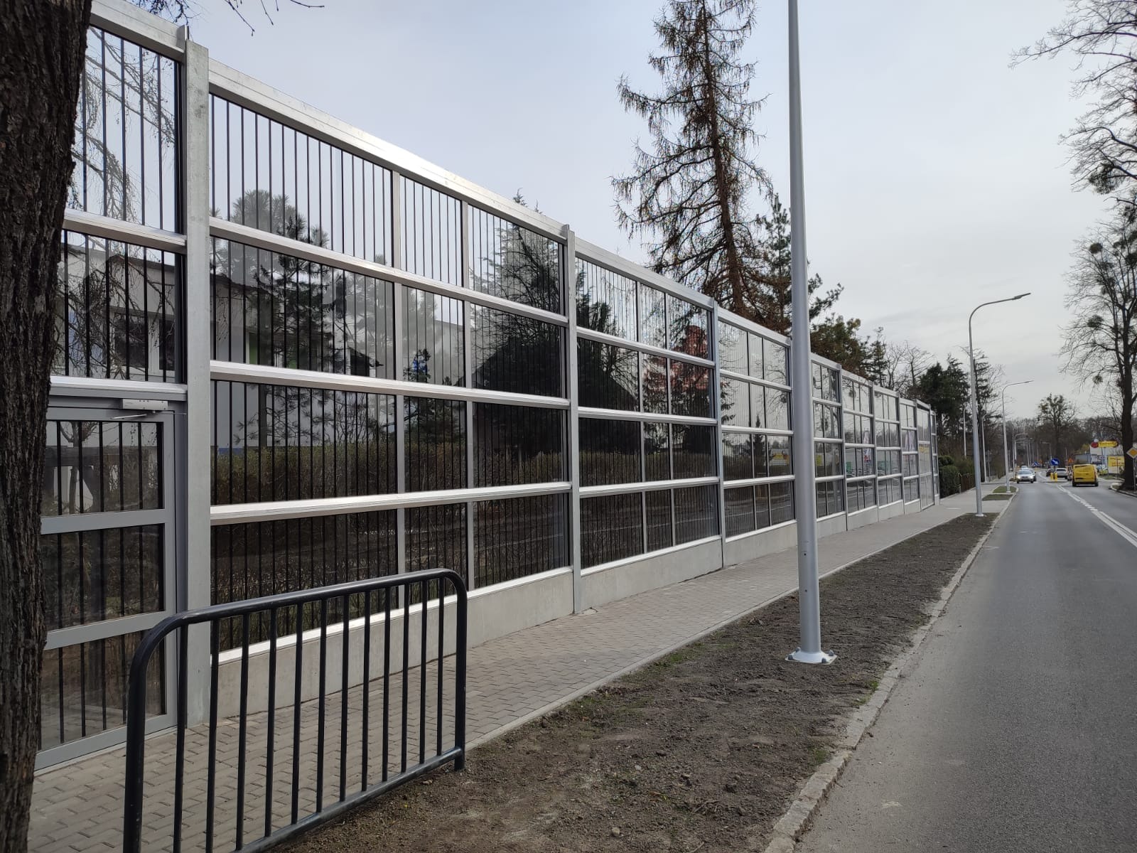 Długa, przezroczysta bariera akustyczna z metalową ramą, oddzielająca chodnik od jezdni, z widocznymi odbiciami drzew i zabudowań w szklanych panelach.