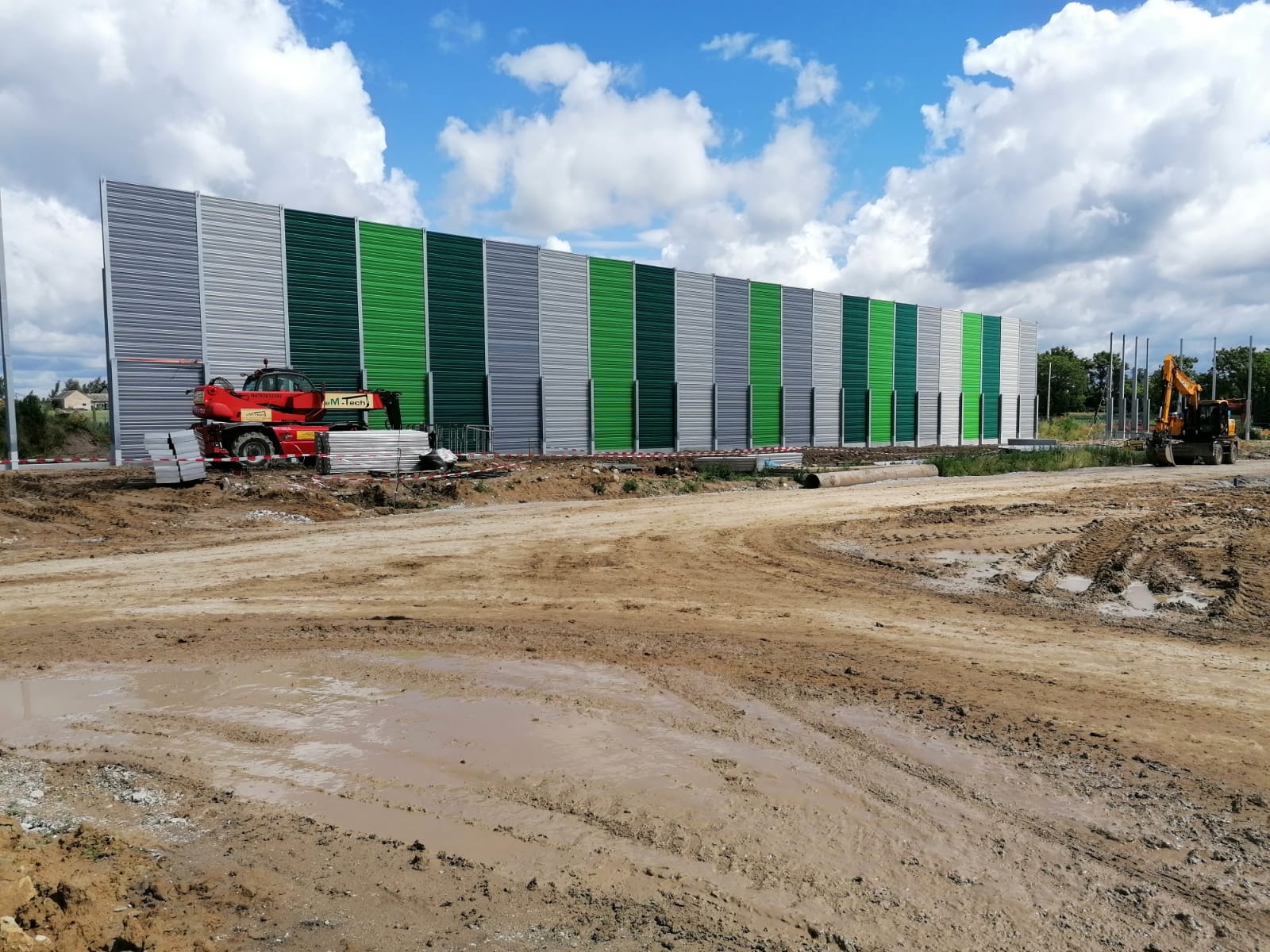 Długi ekran akustyczny w odcieniach szarości i zieleni, w trakcie montażu, z widocznym ciężkim sprzętem budowlanym i błotnistym terenem budowy.