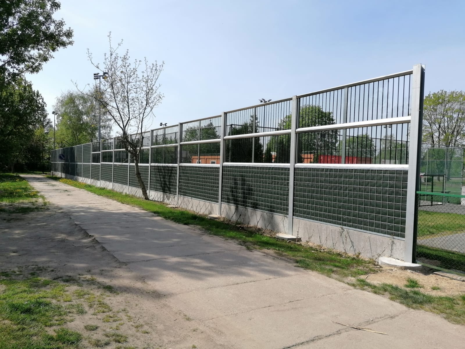 Długi, dźwiękochłonny ekran akustyczny z metalową ramą i wypełnieniem z zielonych paneli w kratkę, oddzielający ścieżkę od terenu sportowego.