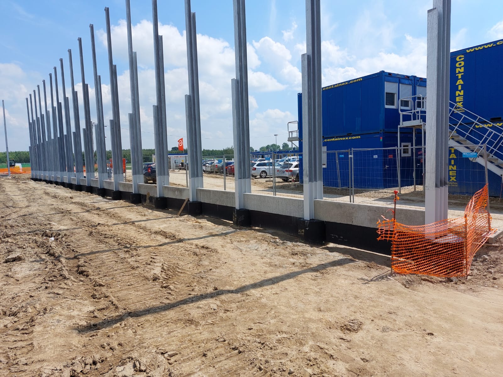 Wznoszenie konstrukcji stalowej: widok na rząd stalowych słupów osadzonych w betonowym fundamencie, zabezpieczonym czarną izolacją, z widocznymi śladami opon na ziemi i kontenerami biurowymi w tle.