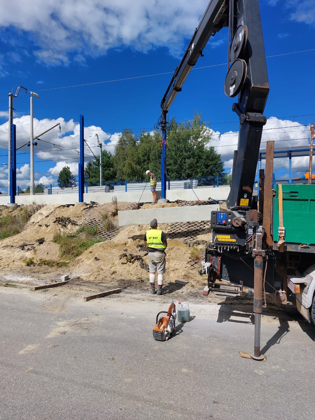 Montaż prefabrykowanych elementów betonowych przy użyciu dźwigu samochodowego, widoczni dwaj pracownicy w kamizelkach odblaskowych, w tle słupy trakcji kolejowej i nasyp ziemny.