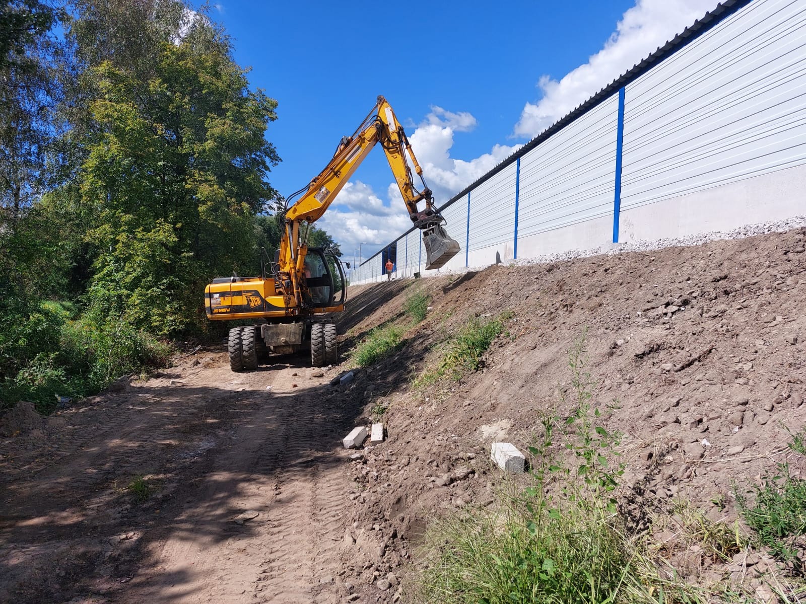 Żółta koparka na nierównym terenie, przygotowująca teren pod budowę, widoczna długa, biała ściana z niebieskimi akcentami w tle.