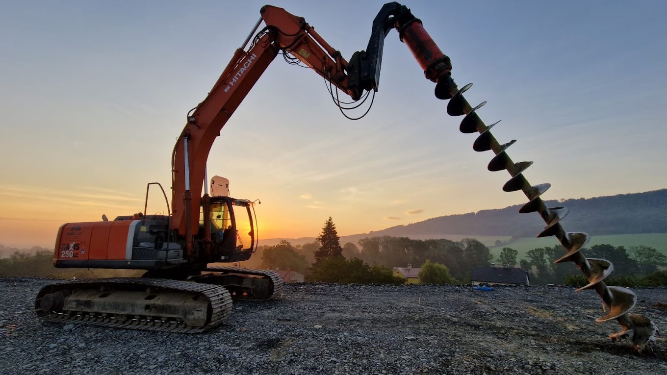 Pomarańczowa koparka Hitachi z wiertłem ślimakowym wzniesiona na tle wschodzącego słońca i zalesionych wzgórz, teren przygotowany pod budowę.
