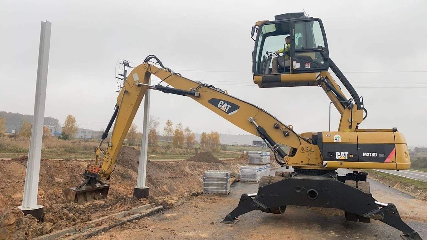 Żółta koparka CAT M318D MM podczas prac ziemnych przy montażu słupów fundamentowych na placu budowy, operator w kabinie.