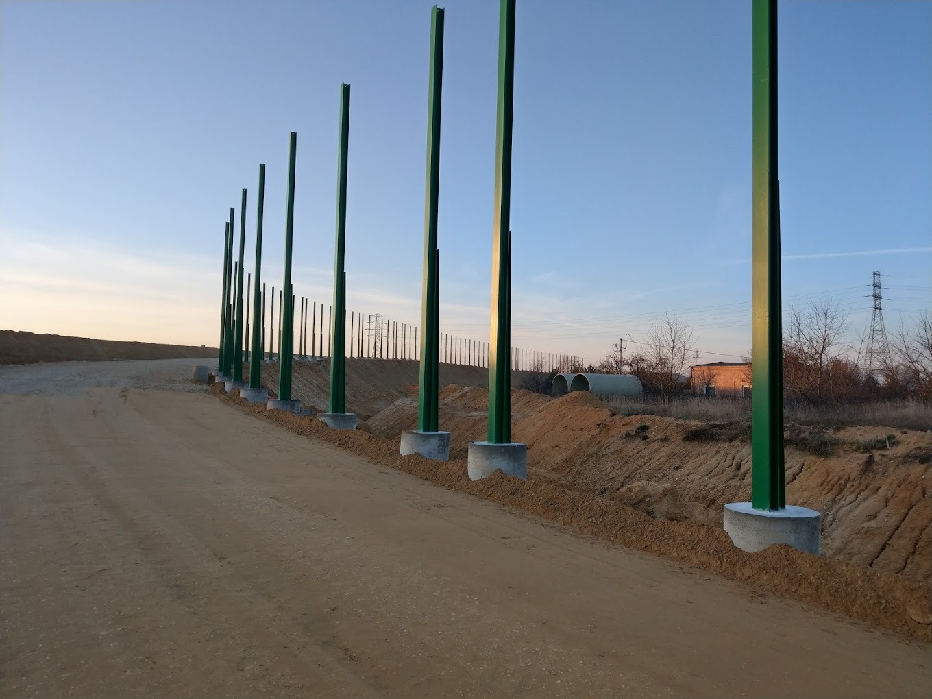 Rząd zielonych słupów konstrukcyjnych osadzonych w betonowych fundamentach, widoczny teren budowy i elementy infrastruktury w tle, wczesny etap budowy.