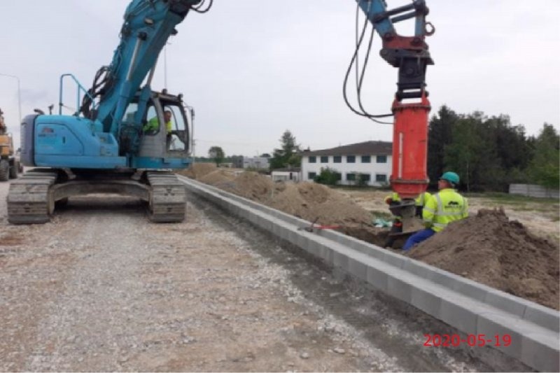 Układanie krawężnika drogowego z użyciem specjalistycznej maszyny w wykopie; w tle koparka i budynek.