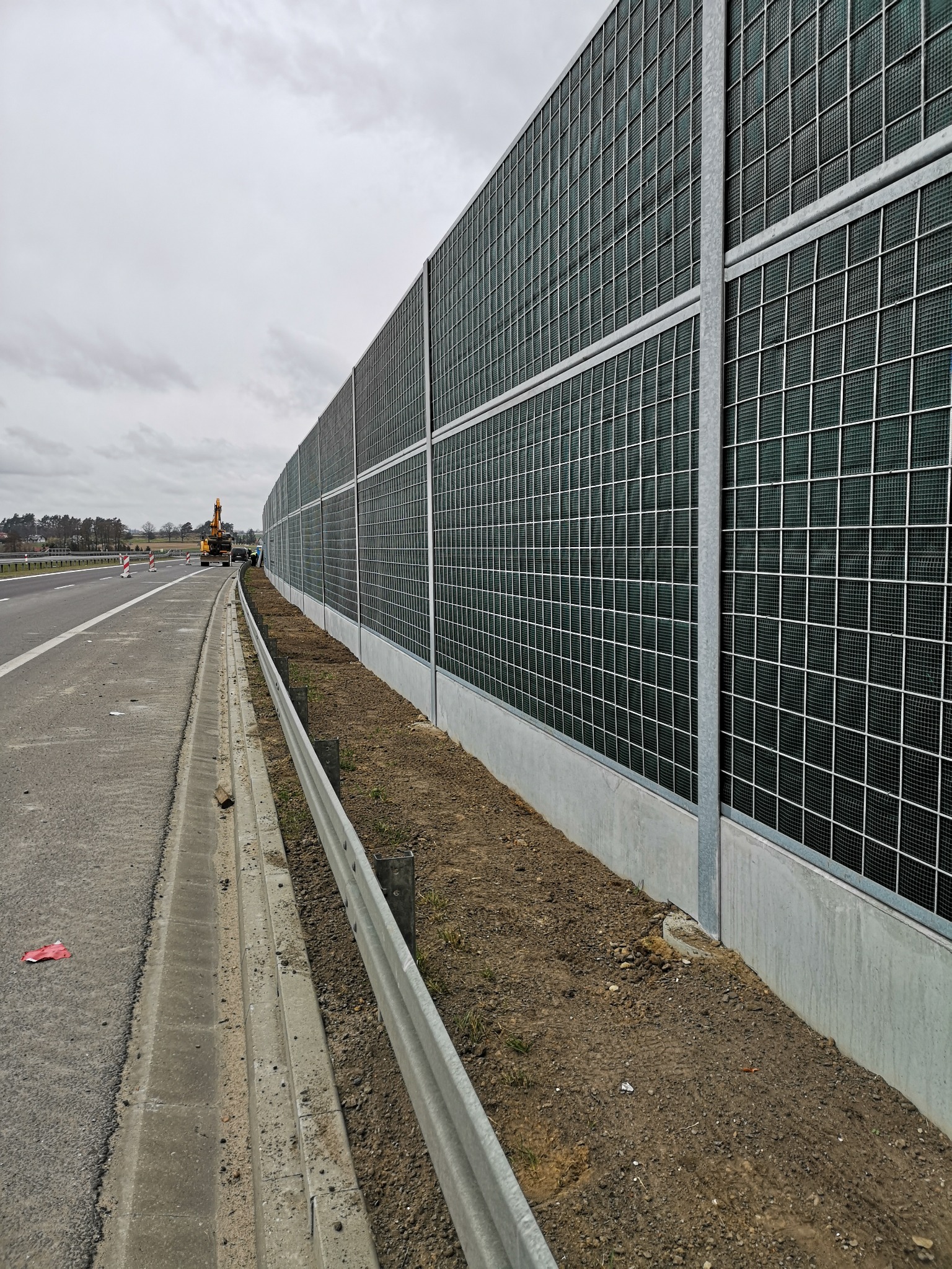 Długa, akustyczna ściana z metalową siatką i betonową podstawą wzdłuż drogi, z koparką w tle i pochmurnym niebem.