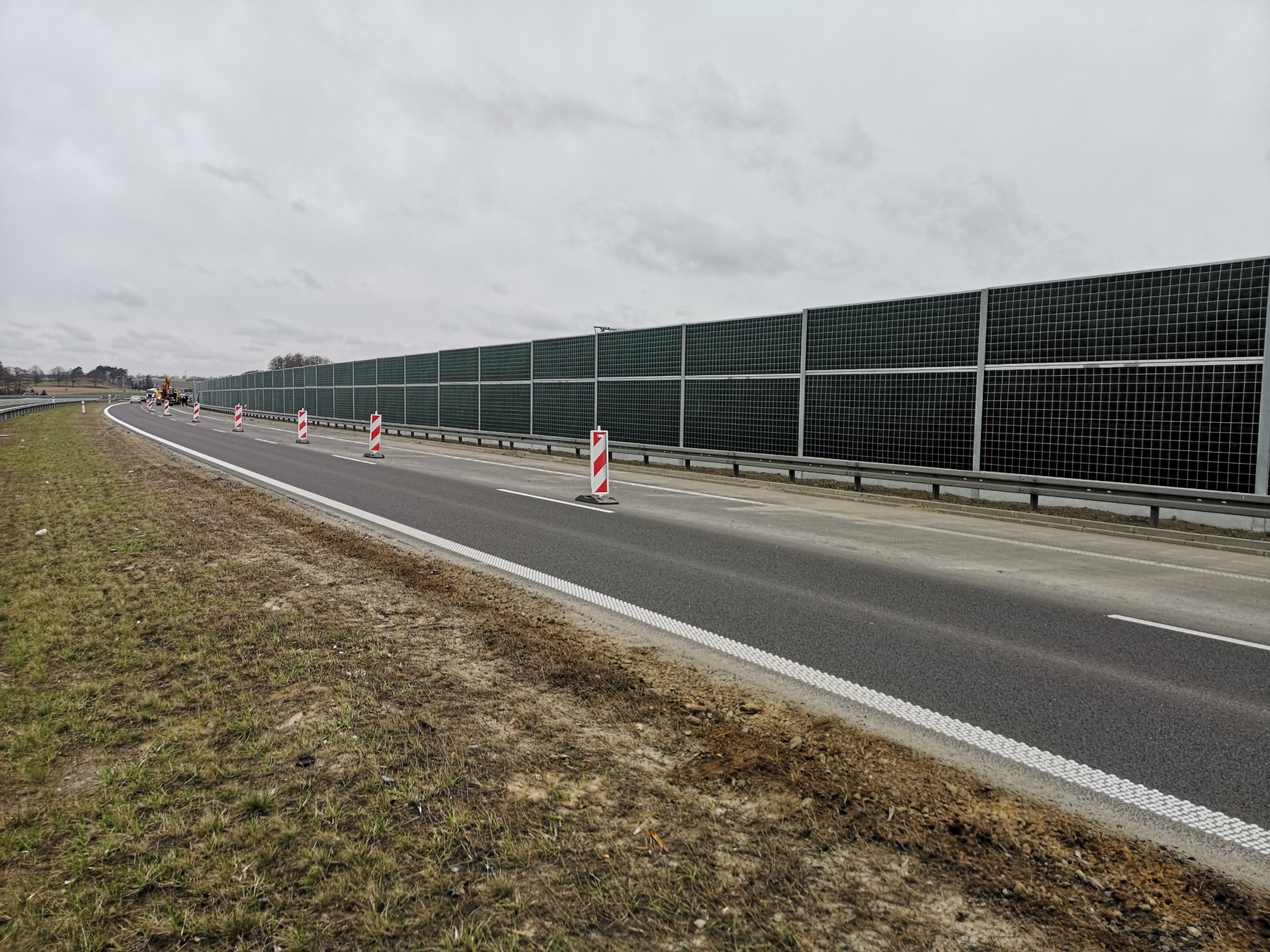 Długa, ciemnozielona bariera akustyczna wzdłuż drogi, zabezpieczona pachołkami z czerwono-białymi pasami, z widoczną koparką w oddali i pochmurnym niebem.
