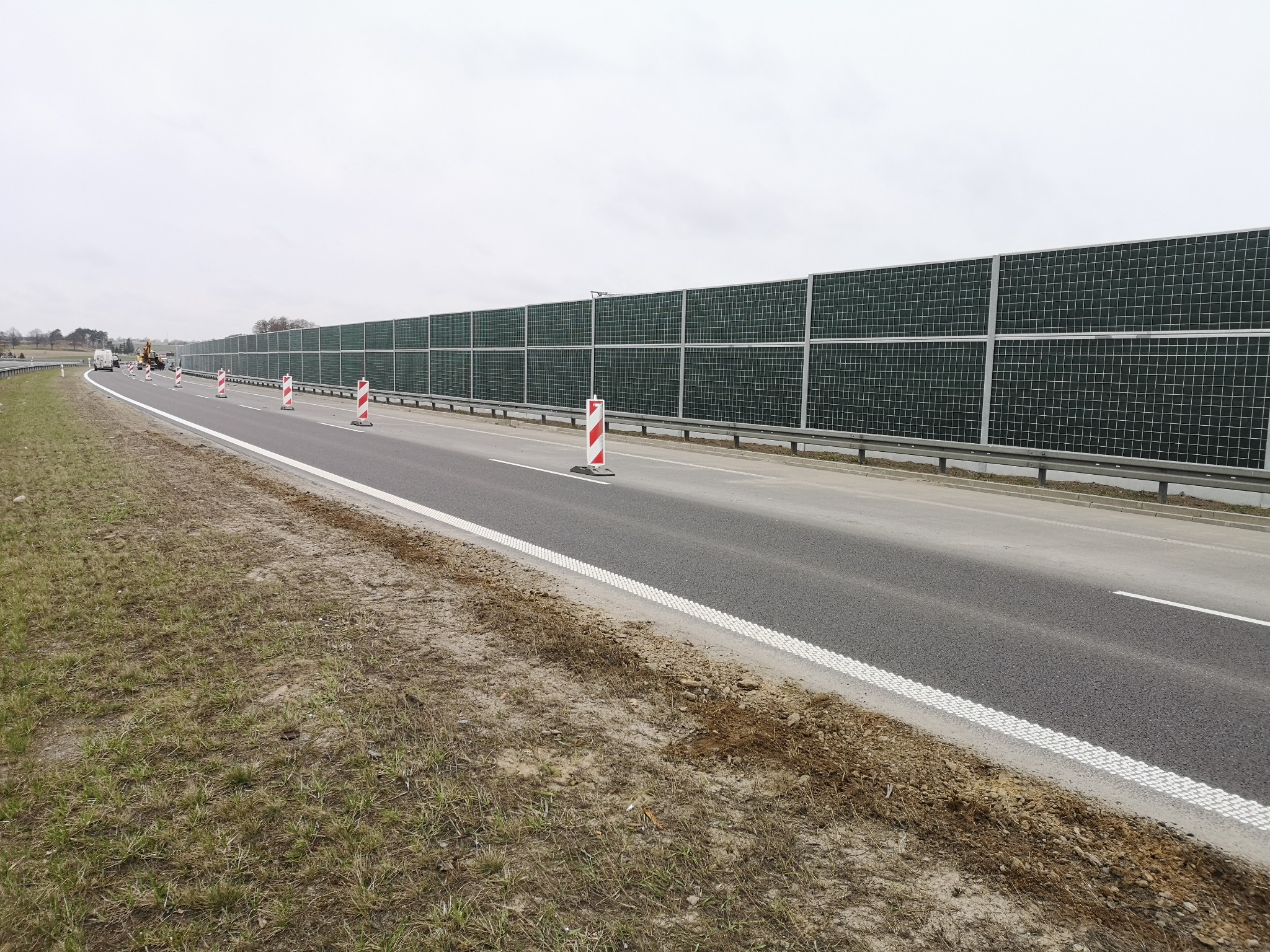 Długa bariera akustyczna z zielonymi panelami wzdłuż drogi, z widocznymi pachołkami ostrzegawczymi i sprzętem budowlanym w oddali.