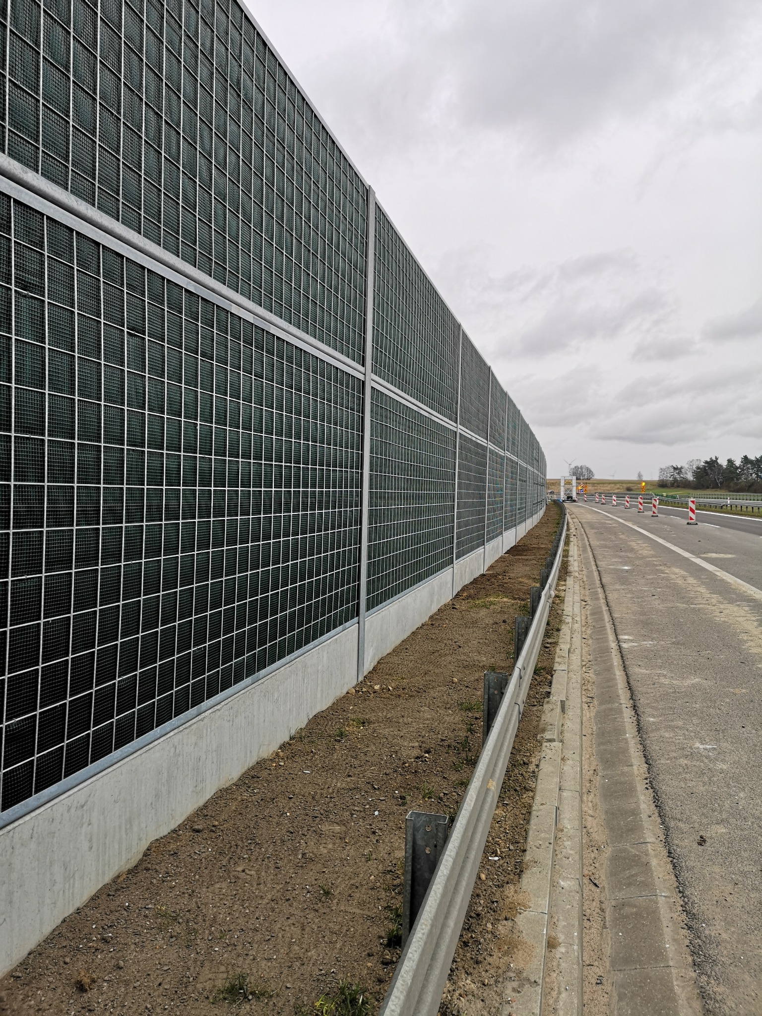 Długa, dźwiękochłonna bariera wzdłuż autostrady, wykonana z paneli z siatki na betonowej podstawie, z widocznymi znakami ostrzegawczymi w oddali.
