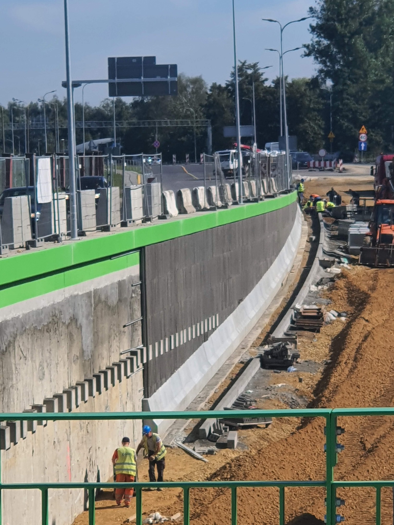 Budowa betonowego muru oporowego w Lusowie, widoczne elementy konstrukcyjne, pracownicy w odblaskowych kamizelkach i ciężki sprzęt budowlany.
