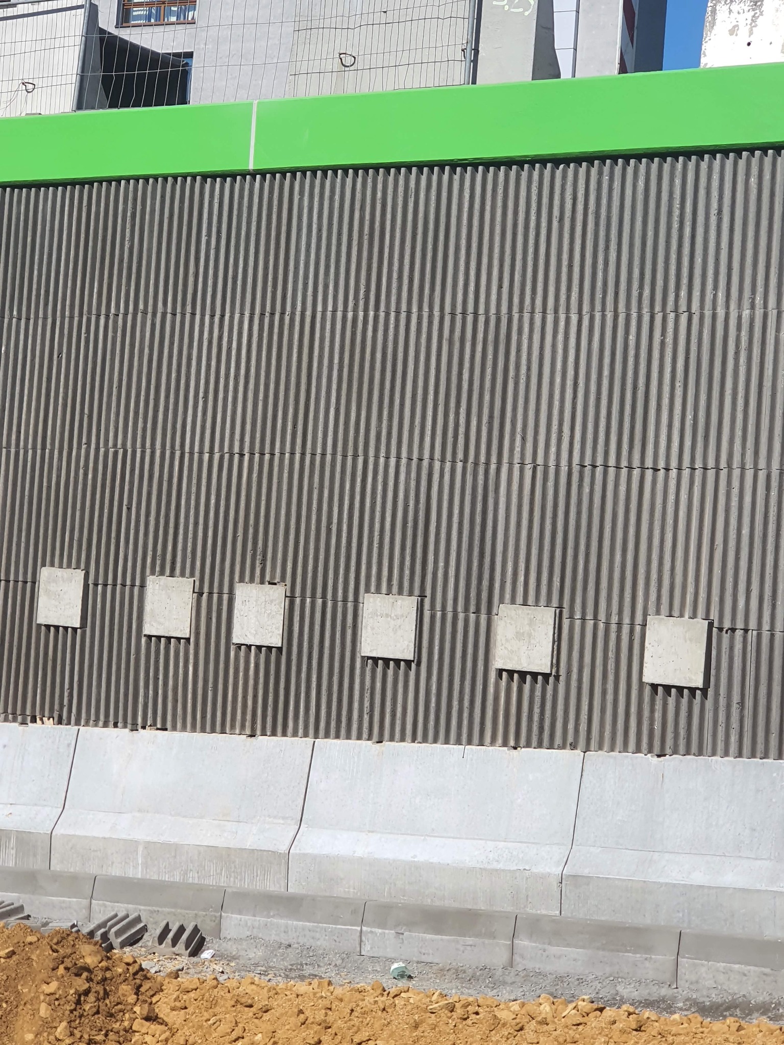 Szara, falista ściana oporowa z betonu z zielonym pasem na górze i pięcioma kwadratowymi elementami mocującymi, widoczna krawężnik i ziemia.
