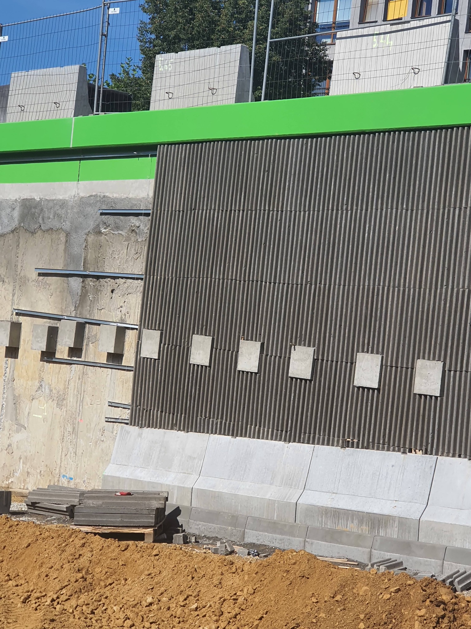 Konstrukcja oporowa z betonu i falistej blachy, wzmocniona metalowymi elementami, widok z dołu na placu budowy z ziemią i paletą betonowych elementów.