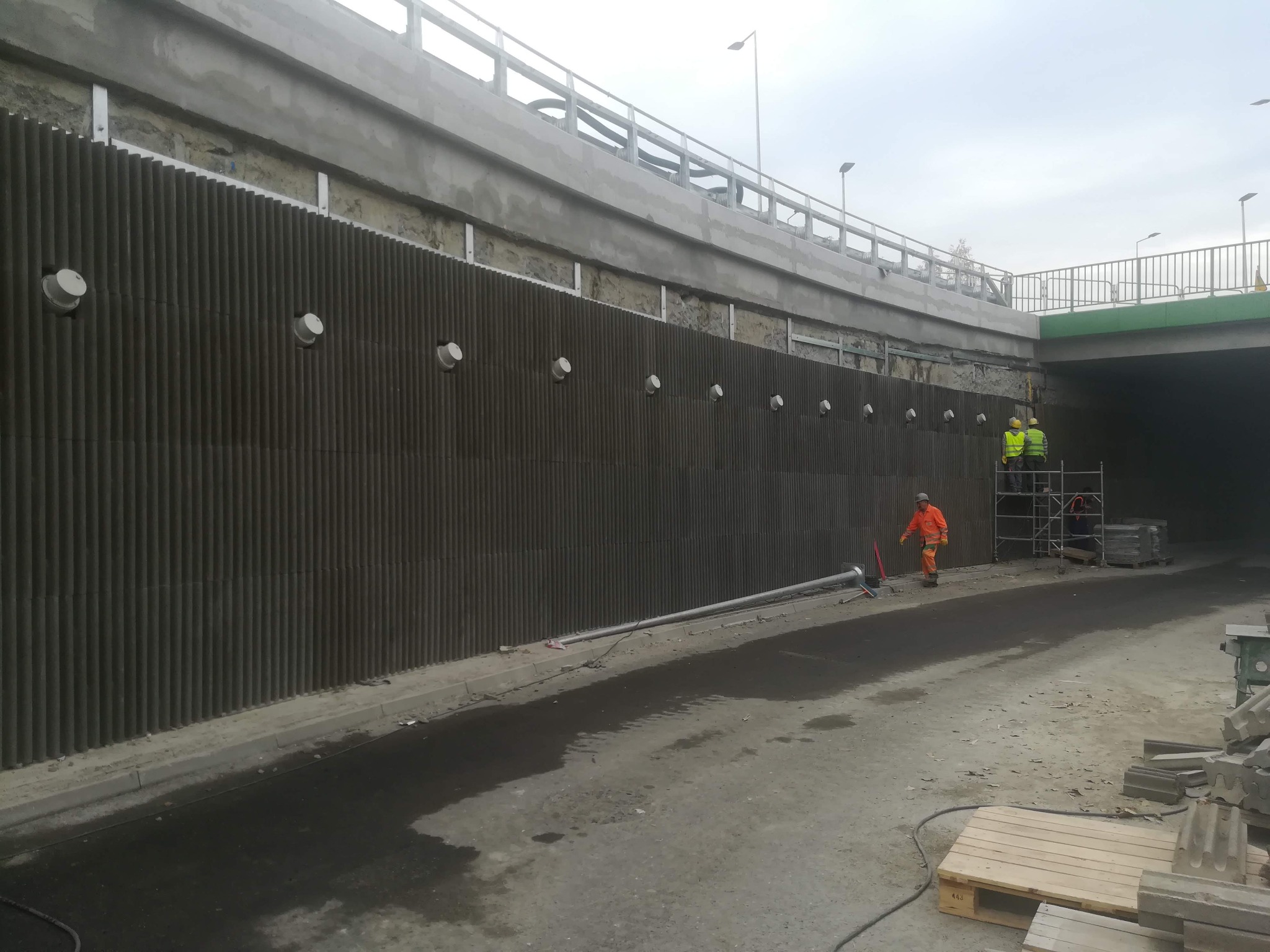 Montaż pionowych paneli akustycznych przy betonowym wiadukcie, pracownicy w odblaskowych ubraniach na rusztowaniu, widoczne mocowania paneli.