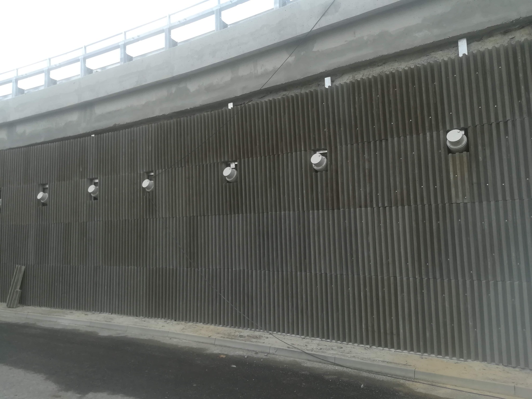 Szara ściana z prefabrykatów betonowych o pionowych żłobieniach, z wbudowanymi okrągłymi otworami wentylacyjnymi i metalowymi wspornikami podtrzymującymi konstrukcję mostu.