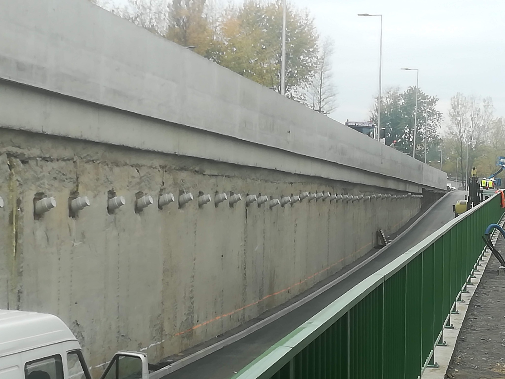 Betonowa ściana oporowa z widocznymi kotwami gruntowymi, zabezpieczająca skarpę przy drodze, widok zza zielonej barierki.