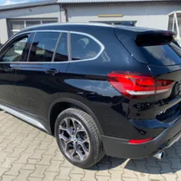 Błyszczący czarny BMW X1 po detailingu, widok z tyłu i boku, felgi aluminiowe, budynek warsztatu w tle.