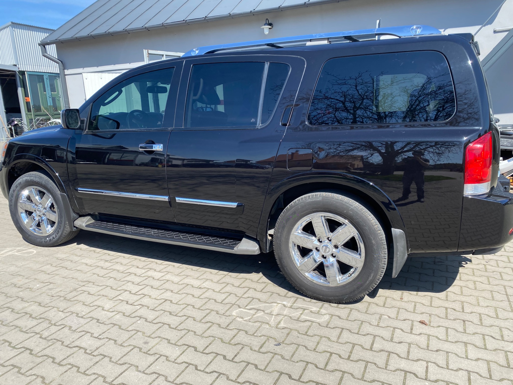 Błyszczący, czarny SUV Nissan Armada z chromowanymi felgami i relingami dachowymi, zaparkowany na brukowanym placu, z odbiciem drzewa i fotografa w szybach.