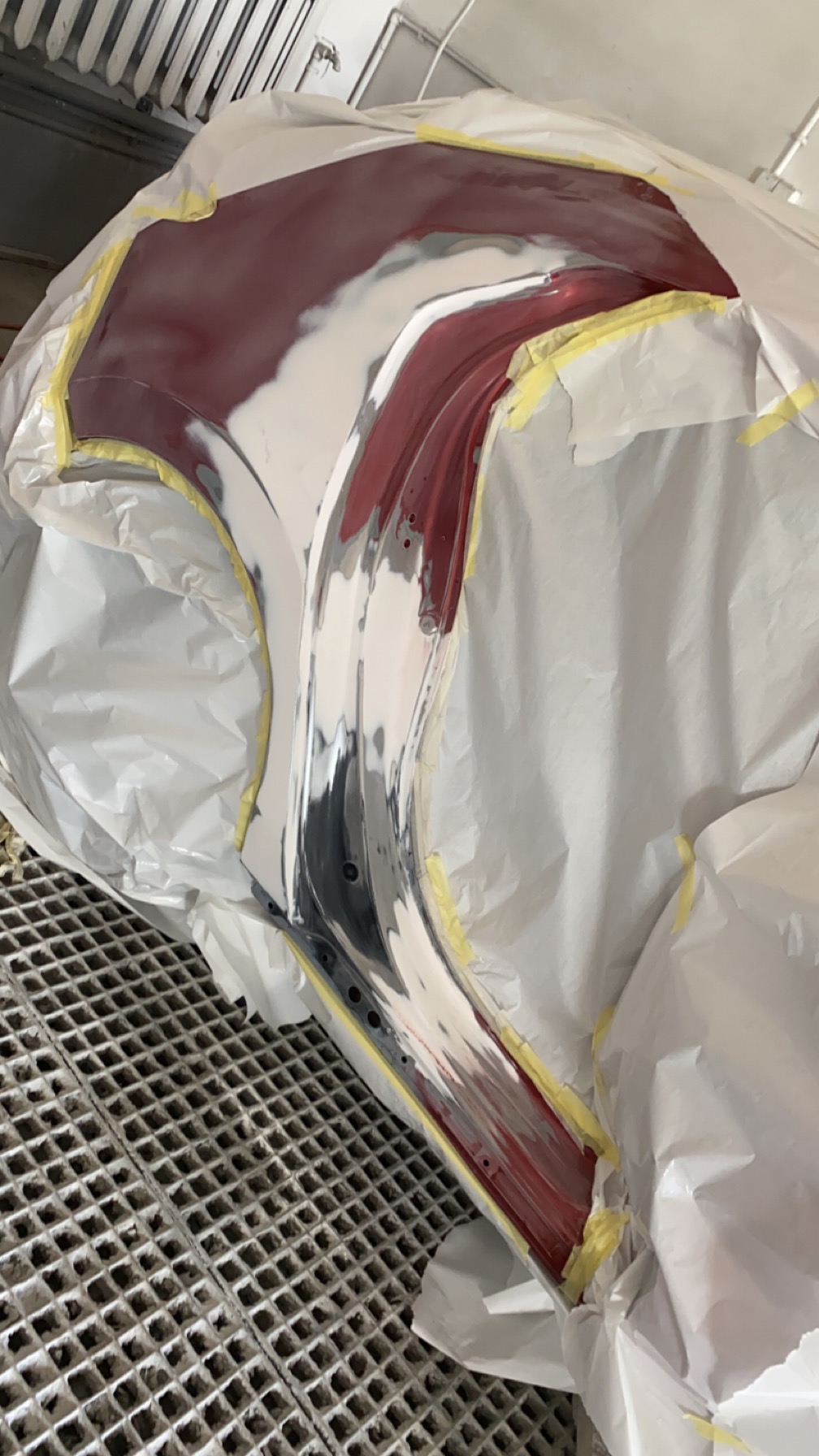 Element karoserii samochodowej w trakcie naprawy lakierniczej, zabezpieczony taśmą i papierem, widoczny częściowo nałożony podkład i czerwona farba, całość na metalowej kratownicy w warsztacie.