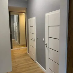 Korytarz w odcieniach szarości z drewnopodobną podłogą, białymi drzwiami z poziomymi frezowaniami i lustrem na końcu, odbijającym kolejne drzwi.