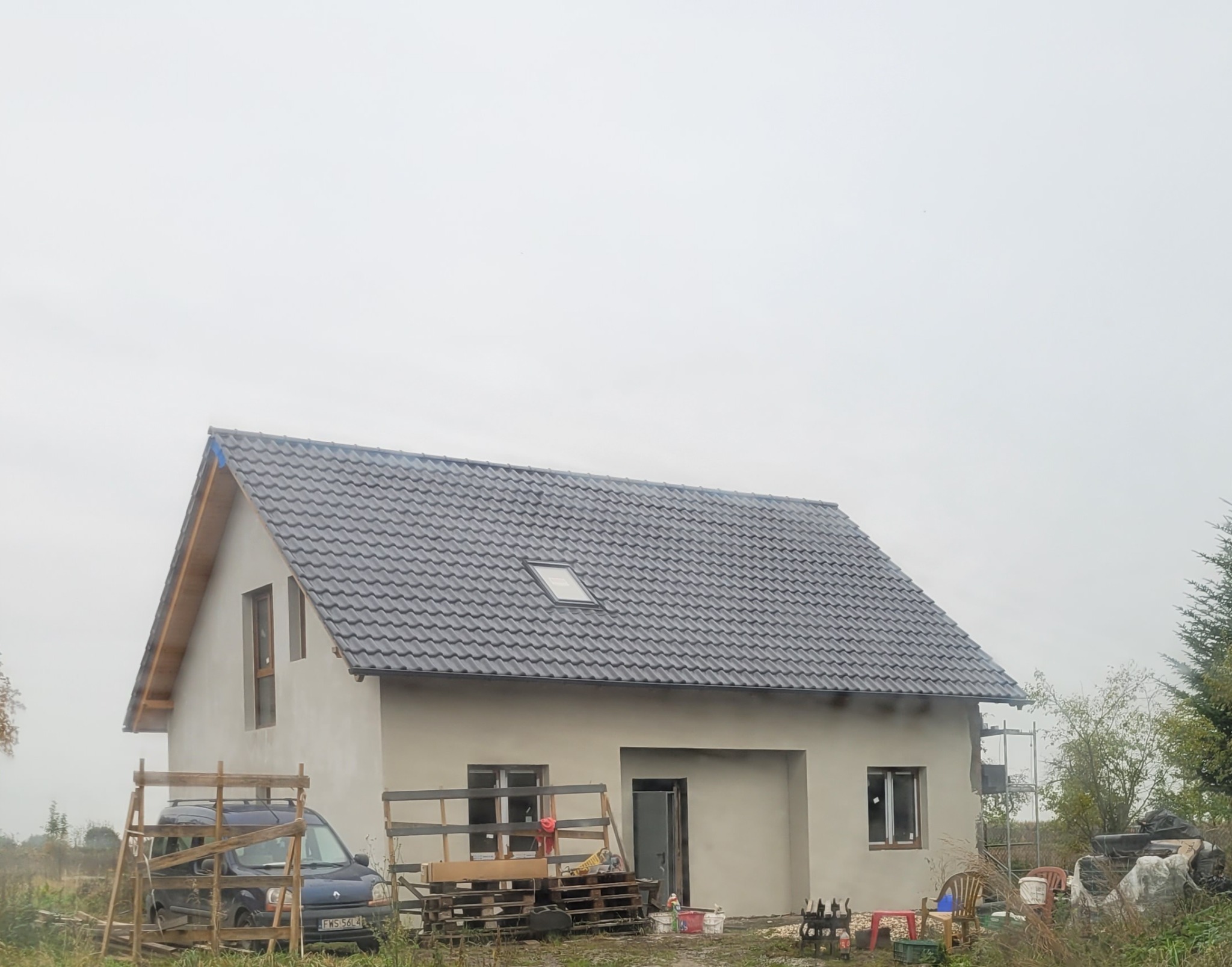 Dom jednorodzinny w trakcie prac wykończeniowych elewacji, widoczne rusztowania, samochód dostawczy oraz palety z materiałami budowlanymi. Dach pokryty ciemną dachówką, okna częściowo zamaskowane...