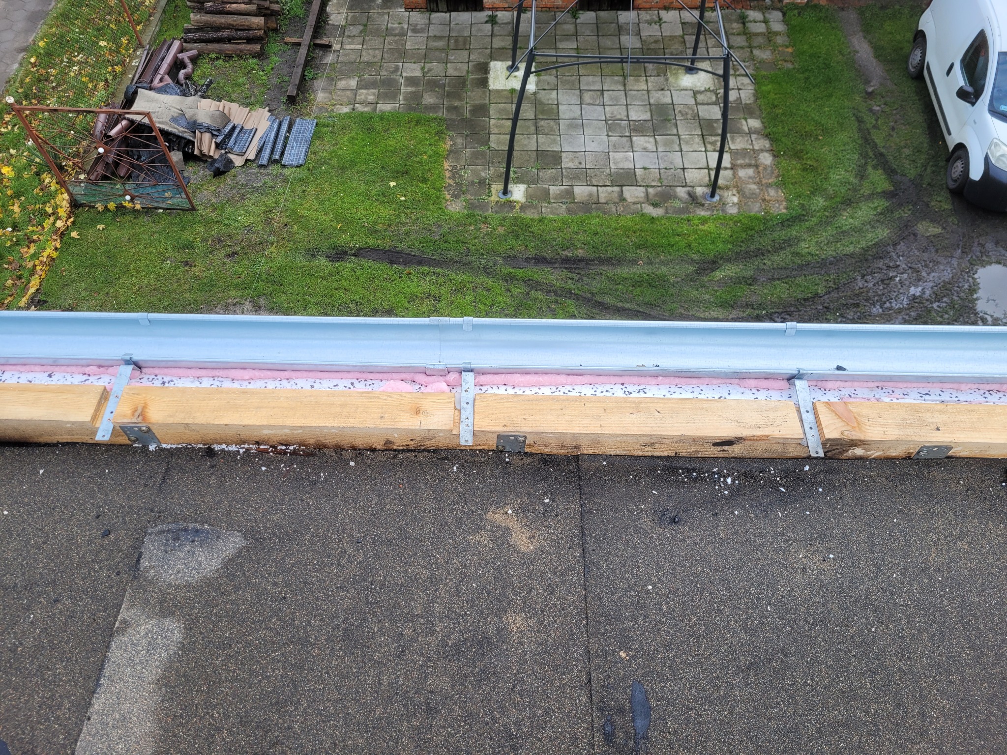 Widok z góry na zamontowany system rynnowy z drewnianą konstrukcją wsporczą, izolacją piankową i metalowymi uchwytami na bitumicznym dachu, z widocznym fragmentem posesji: trawnik, kostka brukowa...