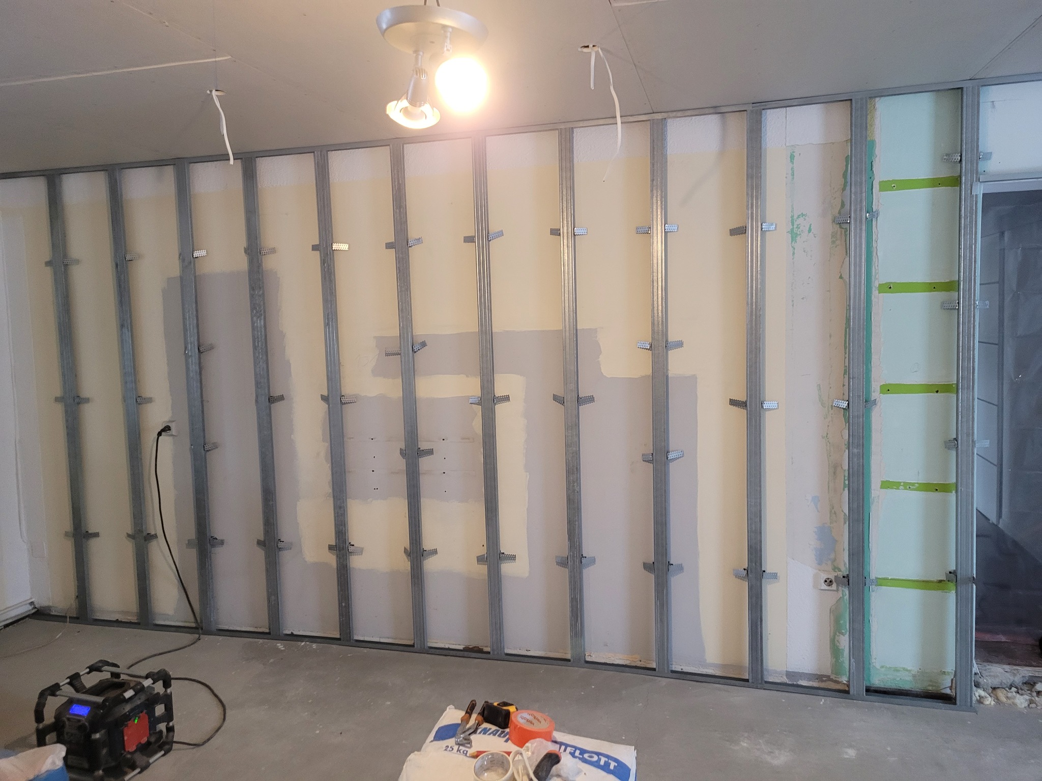 Metalowy stelaż do płyt gipsowo-kartonowych przytwierdzony do ściany, widoczny kabel elektryczny, torba z zaprawą i narzędzia na podłodze.