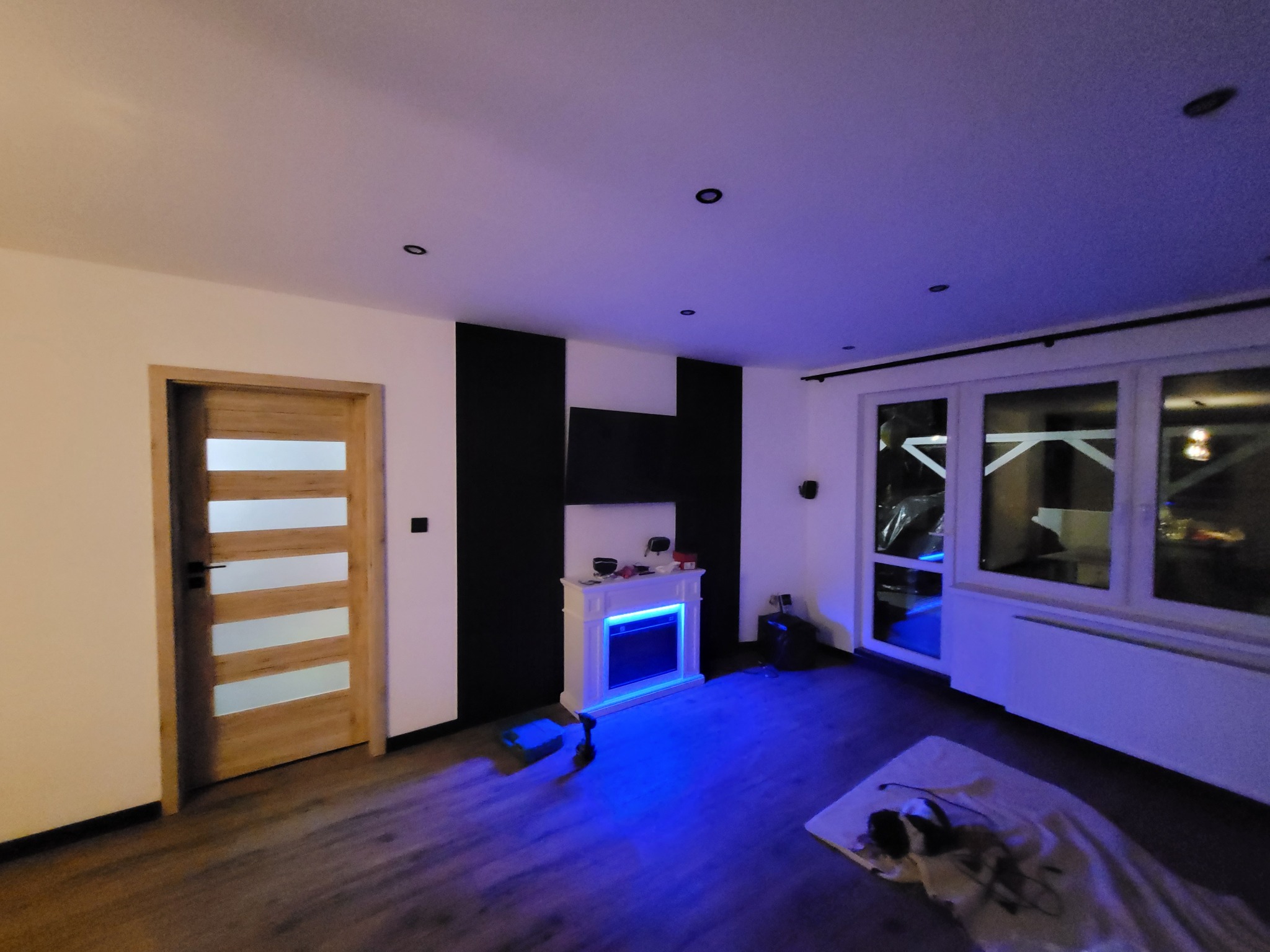 Salon w Nowych Drzewcach z drewnianą podłogą i drzwiami, białą ścianą, czarnym panelem za białą atrapą kominka z niebieskim podświetleniem LED oraz oknem balkonowym.