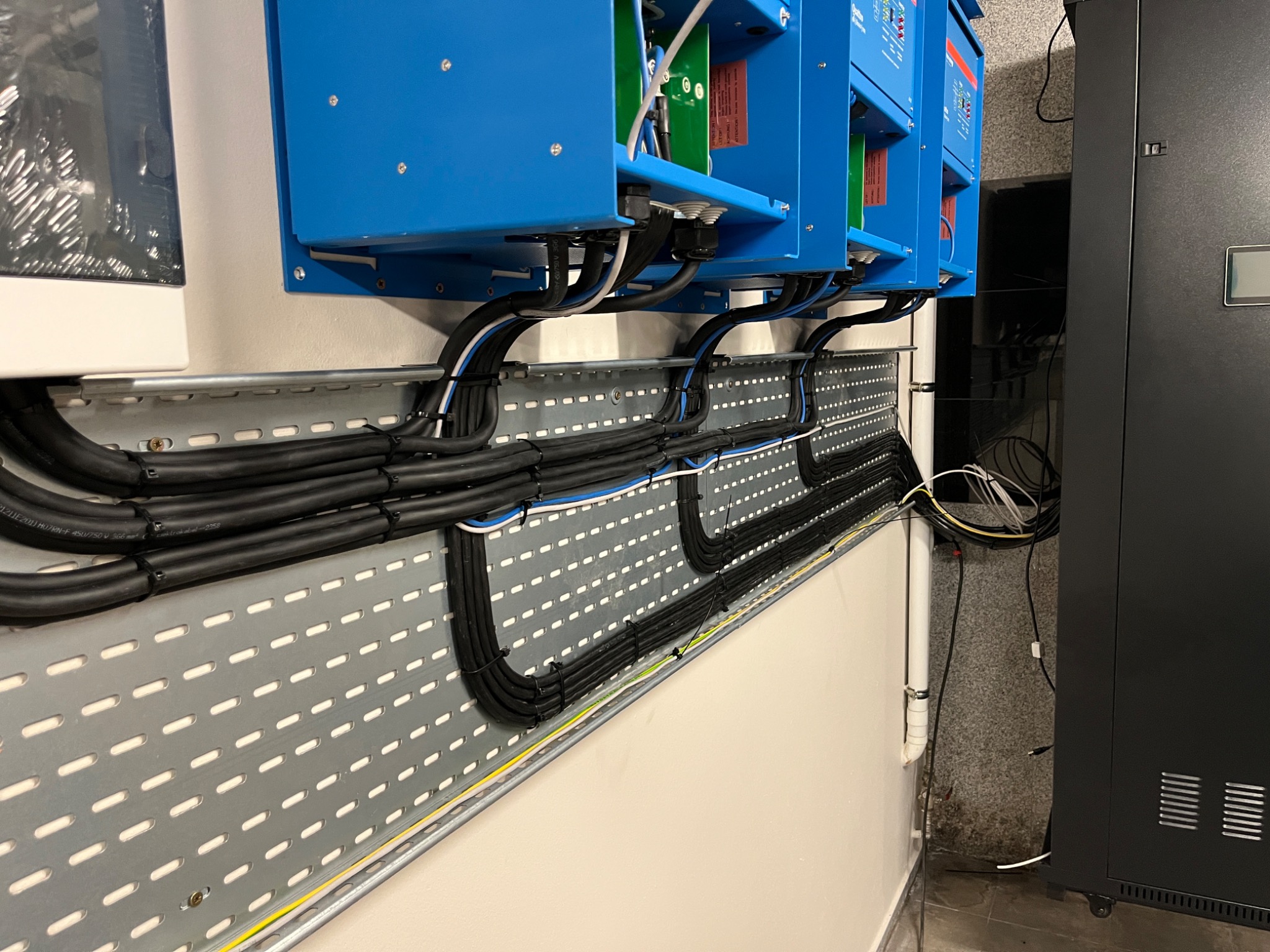 Instalacja elektryczna w Warszawie: Zestaw niebieskich urządzeń elektrycznych zamontowanych na ścianie, z uporządkowanymi kablami prowadzonymi w metalowej korycie kablowej, obok czarnej szafy...