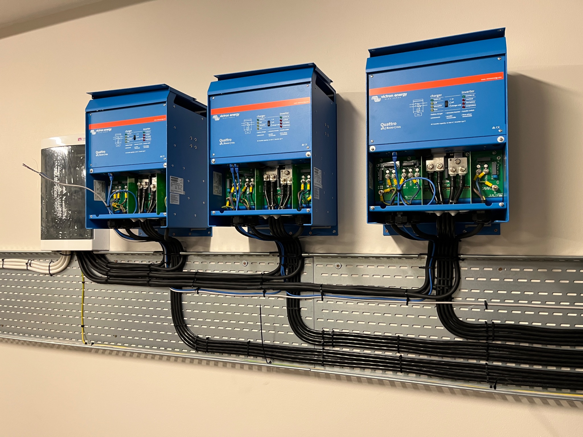 Trzy niebieskie falowniki Victron Quattro zamontowane na ścianie, z widocznymi przewodami w korytkach kablowych, obok biały zbiornik.