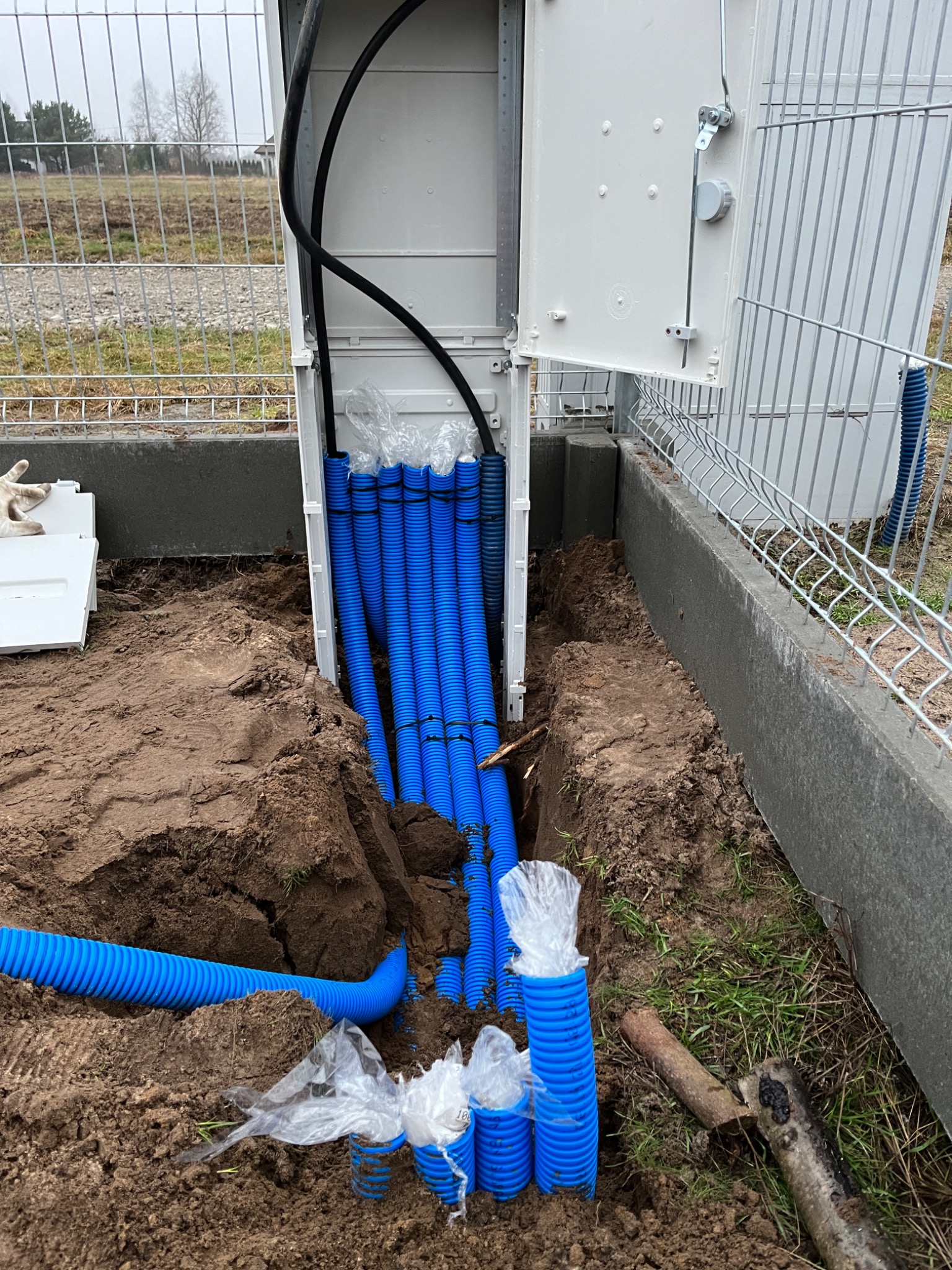 Otwarta skrzynka elektryczna z niebieskimi rurami karbowanymi wychodzącymi z niej i zagrzebanymi w ziemi, widoczne czarne kable wewnątrz skrzynki, teren ogrodzony siatką.