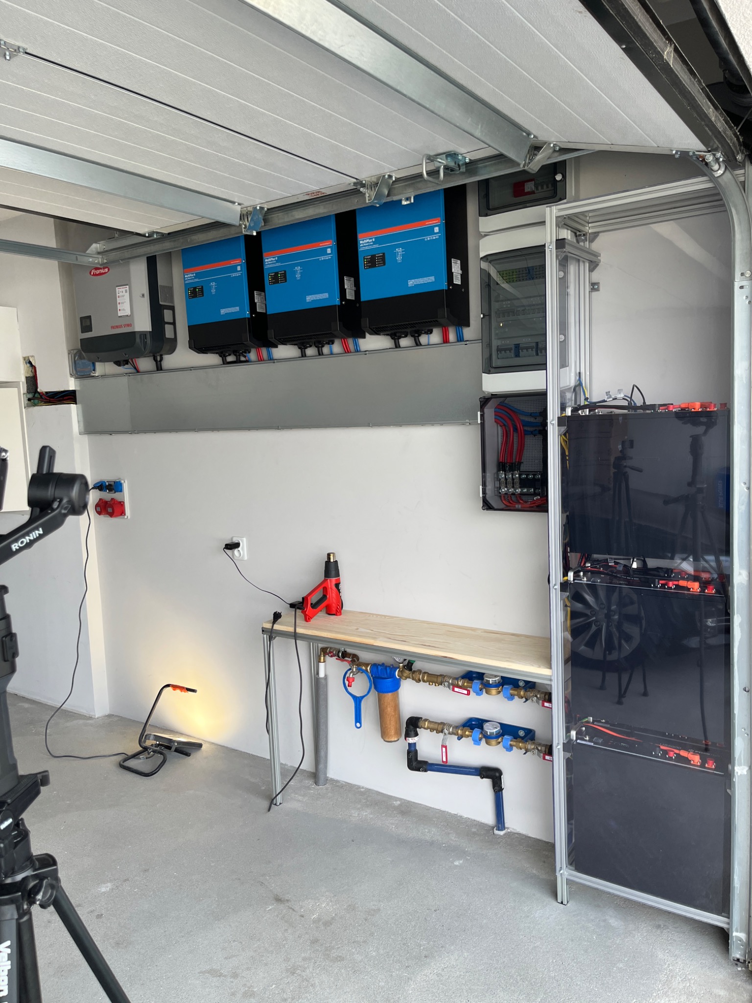 Instalacja fotowoltaiczna w garażu: trzy falowniki, rozdzielnica elektryczna, zestaw akumulatorów, rury hydrauliczne, stół roboczy z opalarką, oświetlenie robocze i statyw kamery.