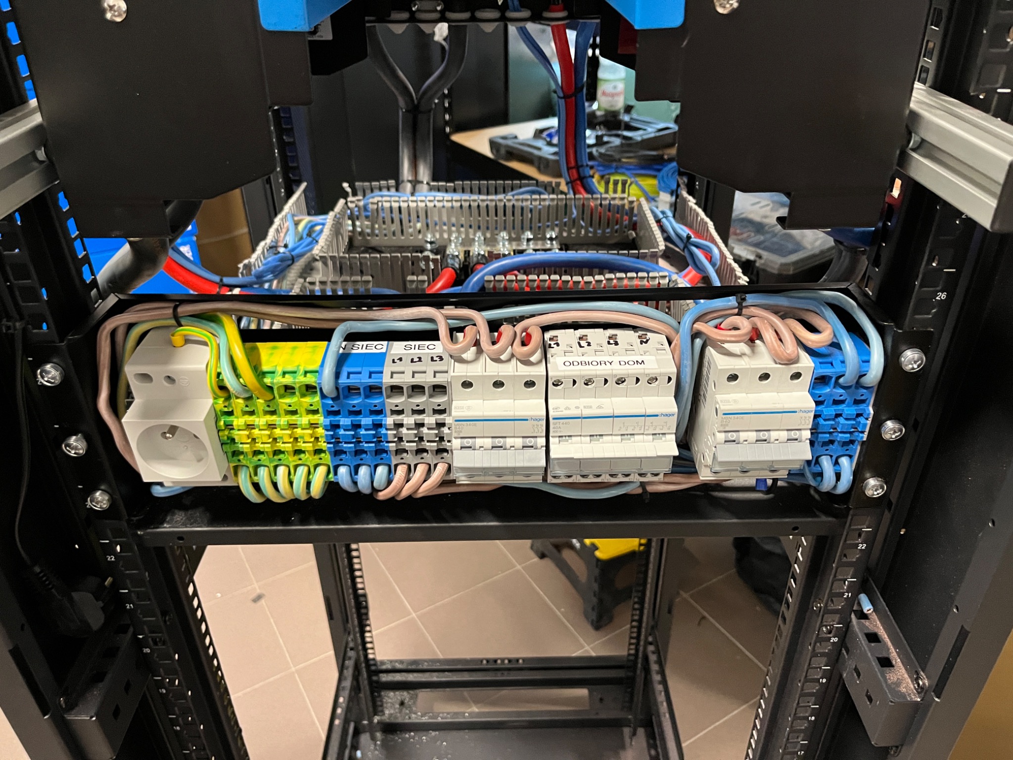 Rozdzielnica elektryczna w szafie rack, widoczne zabezpieczenia, przewody w peszlach, gniazdo elektryczne i listwa zaciskowa.