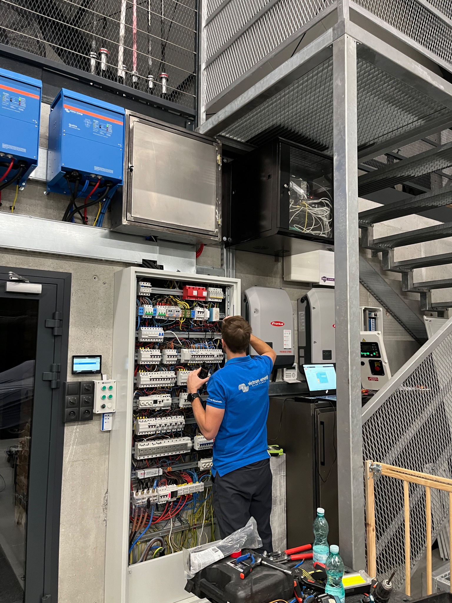 Elektryk w niebieskiej koszulce z logo firmy, stojący przed otwartą szafą elektryczną z licznymi przewodami i bezpiecznikami, w tle widoczne inwertery, schody metalowe i narzędzia na stole.