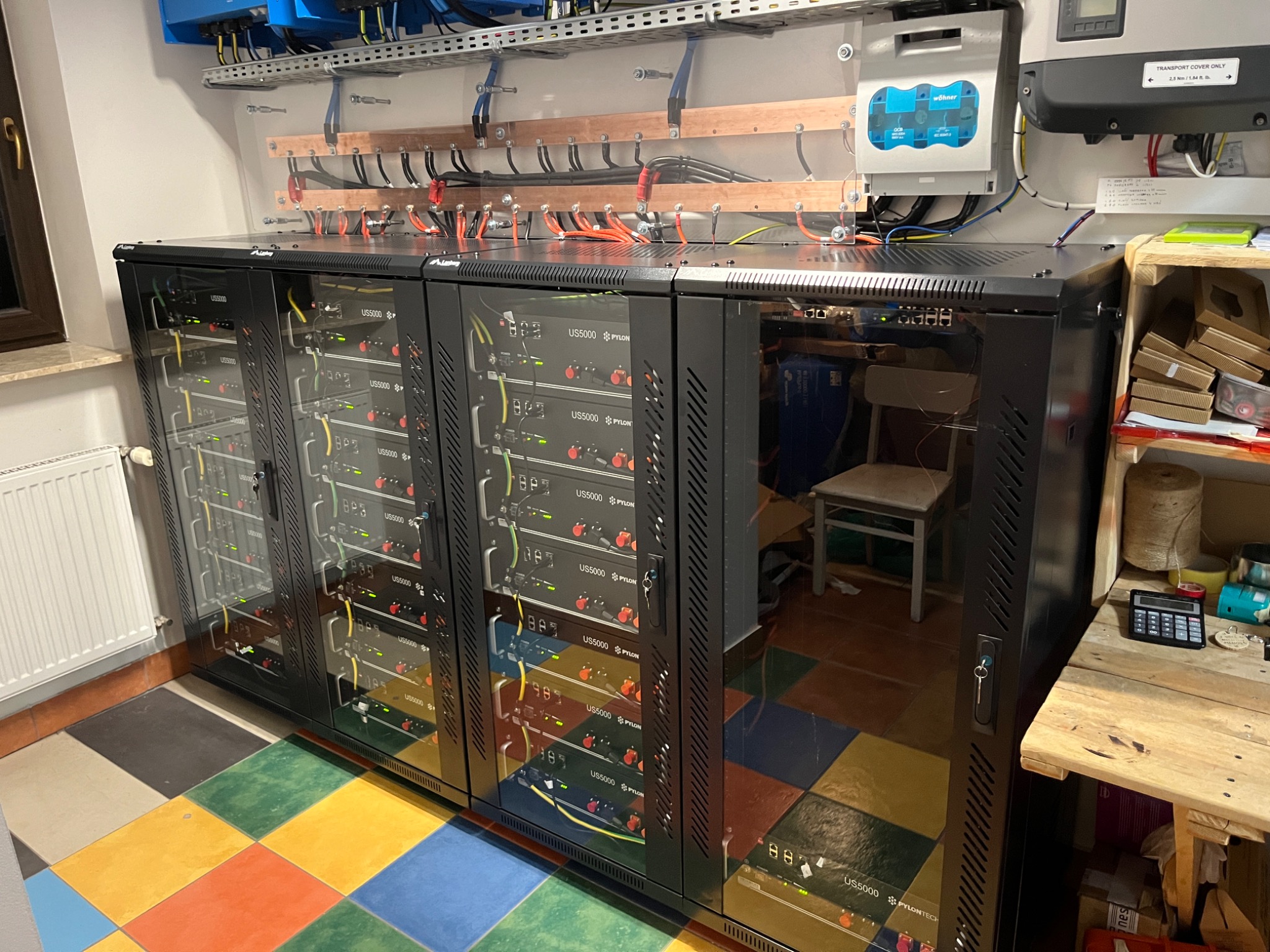 Zestaw akumulatorów Pylontech US5000 w szafach rack, podłączonych do instalacji fotowoltaicznej z widocznymi przewodami i zabezpieczeniami, na tle kolorowej podłogi.