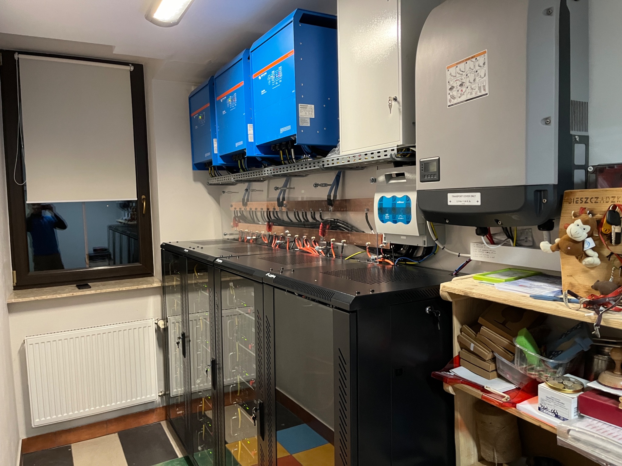 Instalacja fotowoltaiczna w pomieszczeniu: trzy niebieskie falowniki na ścianie, szafa rack z okablowaniem, obok półka z różnymi przedmiotami, widoczne okno z roletą.