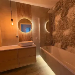 Nowoczesna łazienka z drewnianymi akcentami: okrągłe lustro z podświetleniem LED, umywalka nablatowa, drewniana szafka pod umywalką, wanna z parawanem i tapeta w motywy roślinne.